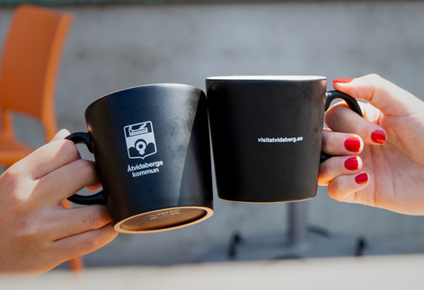 Händer håller kaffekoppar med Åtvidabergs kommuns logotyp på.