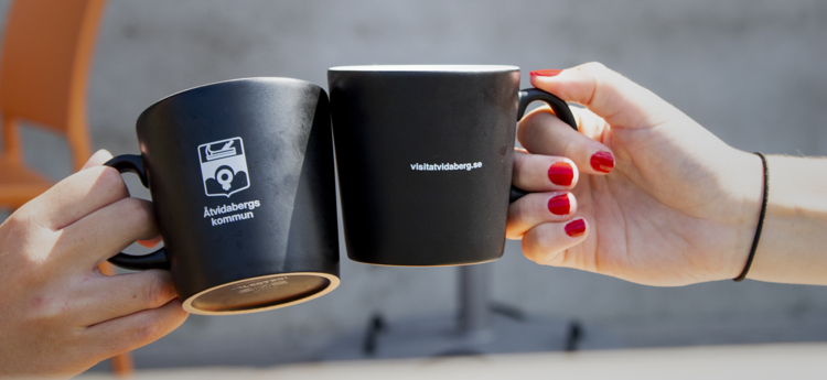 Två händer som skålar med kaffekoppar med Åtvidabergs kommuns logotyp på.